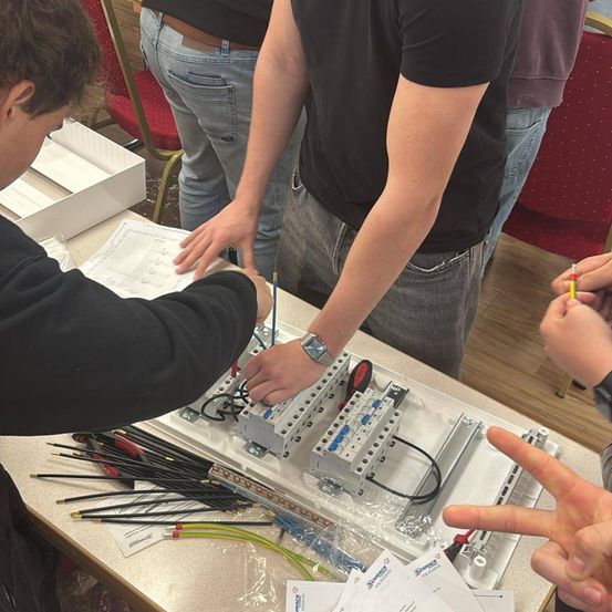 Mehrere Personen arbeiten gemeinsam an einem Stromverteiler, umgeben von verschiedenen Werkzeugen und Kabeln. Papiere sind vor ihnen auf einem Tisch verteilt.