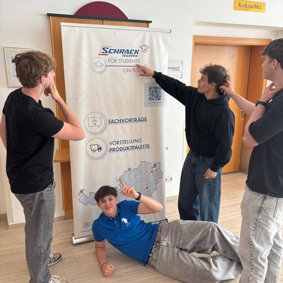 Vier junge Männer stehen um ein Banner herum. Einer zeigt auf einen QR-Code, während die anderen drei stehen. Das Banner sagt 'Schrack Technik für Studenten'.