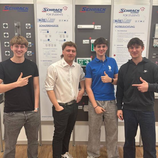 Vier junge Männer stehen vor einer Wand mit elektrischen Installationen. Sie lächeln und zwei von ihnen zeigen den Daumen nach oben. Die Wand hat verschiedene elektrische Komponenten und ein Diagramm.