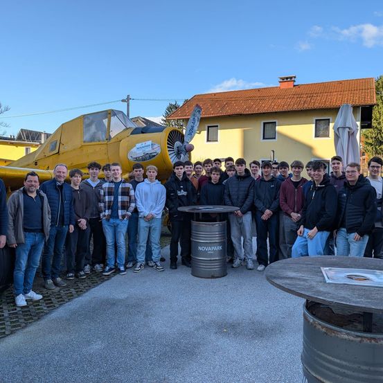 Eine Gruppe von Menschen steht vor einem gelben Flugzeug. Das Flugzeug ist auf dem Boden geparkt und vor ihnen steht ein Fass. Dahinter befindet sich ein Haus mit einem braunen Dach.