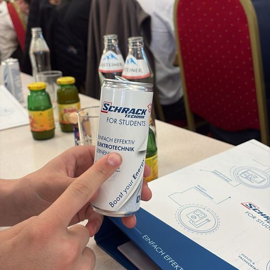 Eine Hand hält eine Dose Energy Drink mit Text für Studenten. Auf dem Tisch stehen verschiedene Getränke und eine offene Mappe.