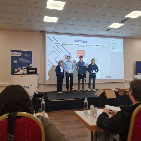 Fünf Personen stehen auf einer Bühne in einem Klassenzimmer mit einer Projektionsleinwand dahinter. Sie halten einen Pokal. Zwei Männer tragen Turnschuhe und zwei tragen Lederschuhe. Eine Frau steht zwischen ihnen.