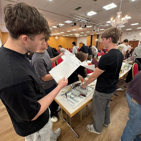 Eine Gruppe junger Männer arbeitet an einer elektrischen Schaltung. Sie stehen um einen Tisch mit Papieren und Werkzeugen herum. Sie scheinen sich in einem Raum mit Holzboden und Kronleuchtern zu befinden.