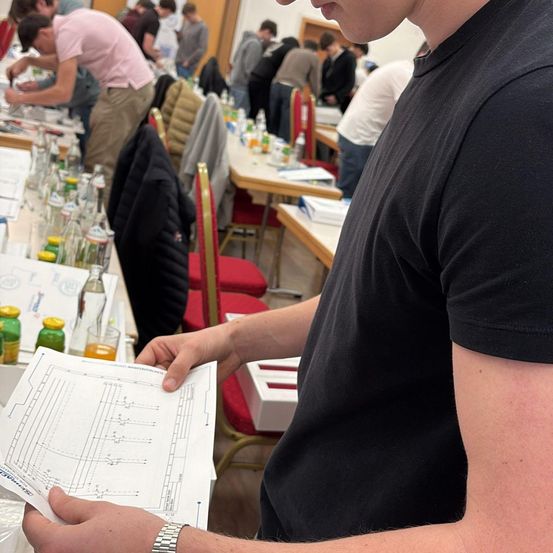 Ein Mann in Schwarz betrachtet einen Diagramm, während andere in einem Raum an einem Tisch mit verschiedenen Flaschen und Papieren arbeiten.