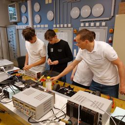 Drei Männer arbeiten in einem Labor mit verschiedenen elektronischen Komponenten und Geräten auf einem Tisch, einschließlich eines Netzteils und einer Flasche. Ein Mann zeigt auf das Netzteil.