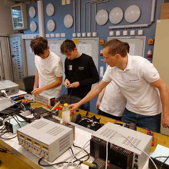 Drei Männer arbeiten in einem Labor mit verschiedenen elektronischen Komponenten und Geräten auf einem Tisch, einschließlich eines Netzteils und einer Flasche. Ein Mann zeigt auf das Netzteil.