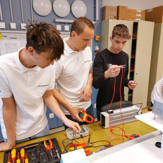 Drei junge Männer arbeiten in einer Werkstatt an einem Projekt mit Werkzeugen und elektrischen Geräten. Einer hält ein Multimeter.