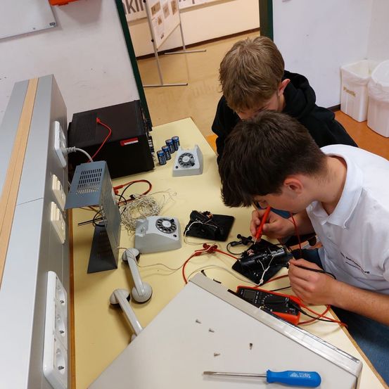 Zwei junge Männer arbeiten an elektronischen Geräten, wahrscheinlich in einem Klassenzimmer. Einer lötet, während der andere zusieht. Verschiedene Werkzeuge und Geräte stehen auf einem gelben Tisch.