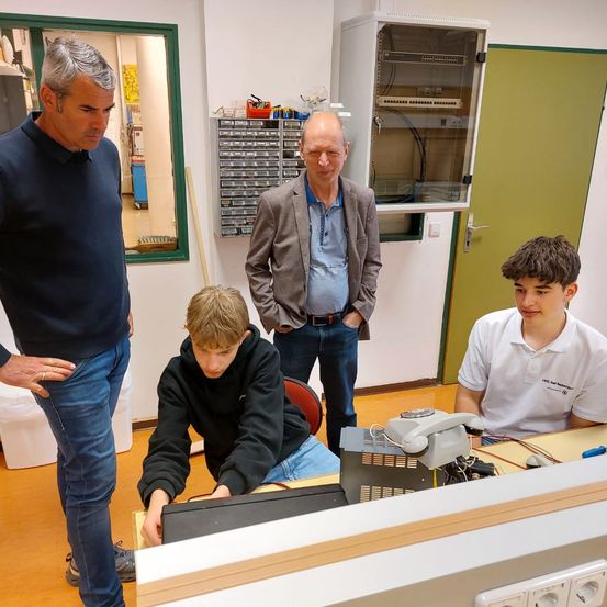 Drei Männer in einer Werkstatt mit einem jungen Jungen, der an einem Computer arbeitet. Ein Mann schaut auf den Computer, während die anderen beiden beobachten.