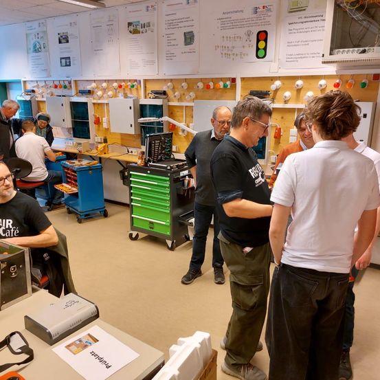 Eine Gruppe von Menschen in einer Werkstatt, die sich im Gespräch befindet. Mehrere Menschen stehen und unterhalten sich, während andere sitzen. Verschiedene Geräte und Werkzeuge sind auf den Tischen und Wänden sichtbar.