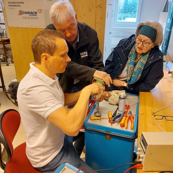 Ein Mann arbeitet mit einem älteren Mann und einer älteren Frau an einem elektrischen Projekt. Sie sind in einem Raum mit Werkzeug auf einem Tisch.