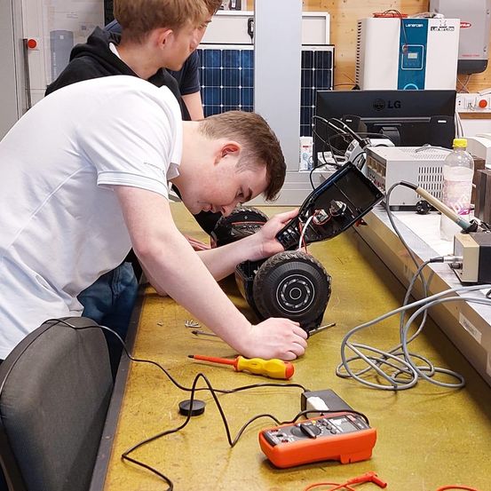Zwei Männer in einer Werkstatt, einer benutzt einen Schraubendreher an einem Gerät, während der andere zusieht. Verschiedene elektronische Komponenten und Werkzeuge sind auf dem Tisch.