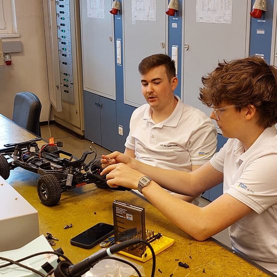 Zwei junge Männer in weißen Poloshirts arbeiten an einem Spielzeugauto auf einem Schreibtisch, einer trägt eine Uhr. Ein Mikrofon steht vor ihnen.