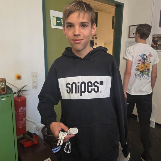 Ein junger Junge in einer Snipes-Kapuzenjacke steht mit Schlüsseln in der Hand lächelnd in der Nähe eines Feuerlöschers. Ein weiterer Mann in einem weißen T-Shirt geht im Hintergrund.