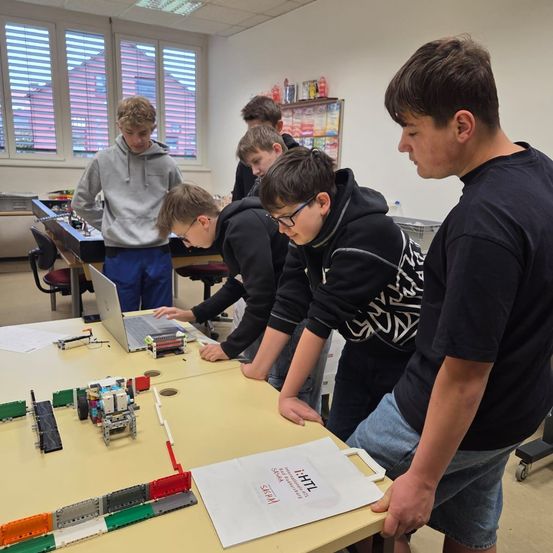 Mehrere junge Schüler arbeiten in einem Klassenzimmer an einem Projekt. Sie sind um einen Tisch versammelt, auf dem ein Laptop und verschiedene Bauklötze stehen. Ein Schüler konzentriert sich auf den Laptop, während andere zuschauen.