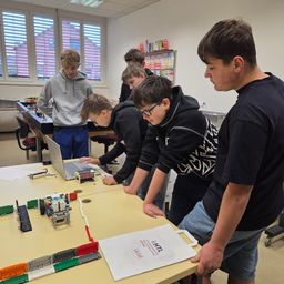 Mehrere junge Schüler arbeiten in einem Klassenzimmer an einem Projekt. Sie sind um einen Tisch versammelt, auf dem ein Laptop und verschiedene Bauklötze stehen. Ein Schüler konzentriert sich auf den Laptop, während andere zuschauen.