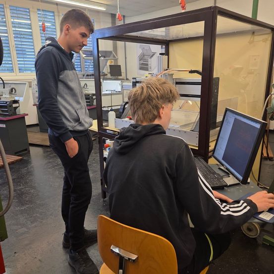 Zwei junge Männer arbeiten in einem Labor an einem Computer, einer schaut auf einen Bildschirm, während der andere steht.