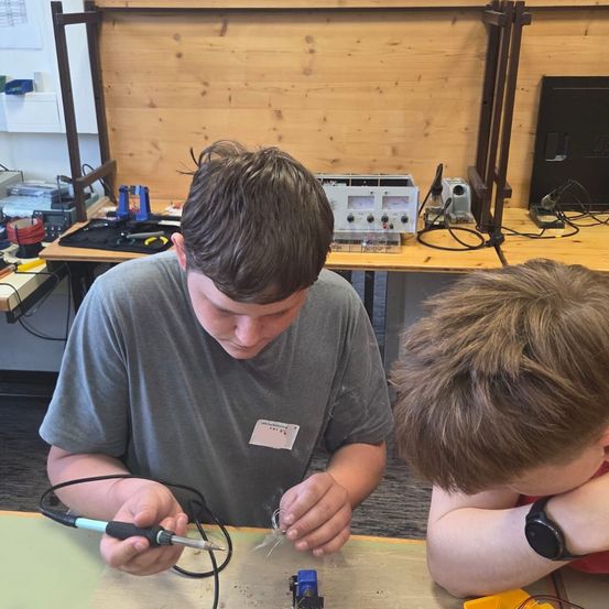 Zwei Jungen sind in einer Werkstatt, einer lötet ein kleines elektronisches Schaltkreis. Ein Tisch mit verschiedenen Werkzeugen und Ausrüstung steht hinter ihnen.