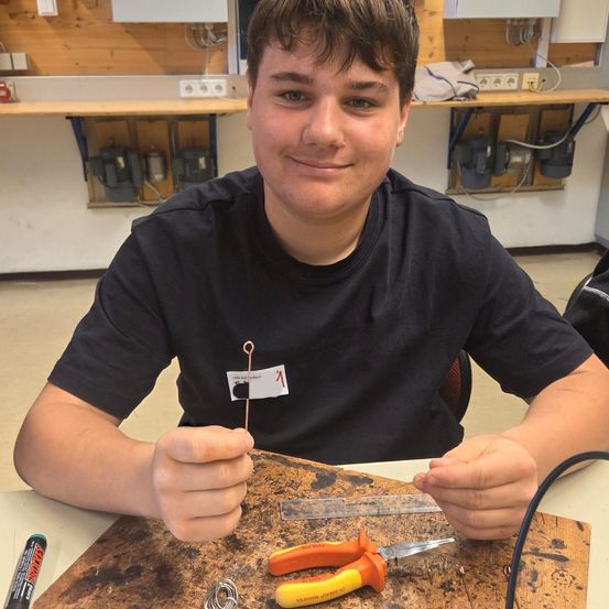 Ein junger Mann arbeitet an einer Holzplatte mit Werkzeugen und Drähten. Er trägt ein schwarzes Shirt und hat einen ernsten Gesichtsausdruck.