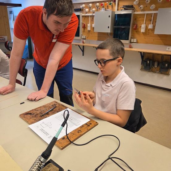 Ein Mann in einem orangefarbenen Shirt hilft einem Jungen mit Brille bei einem Elektronikprojekt in einer Werkstatt. Sie sitzen an einem Tisch mit einem Lötkolben und anderen Werkzeugen.