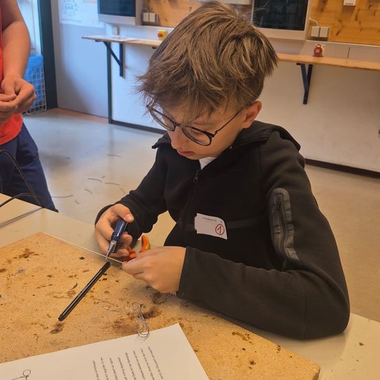 Ein Junge in einer Werkstatt, mit Werkzeugen, sitzt an einem Schreibtisch. Er trägt eine Brille und einen schwarzen Pullover. Er arbeitet an einem Projekt und hat ein Blatt Papier mit Anweisungen vor sich.