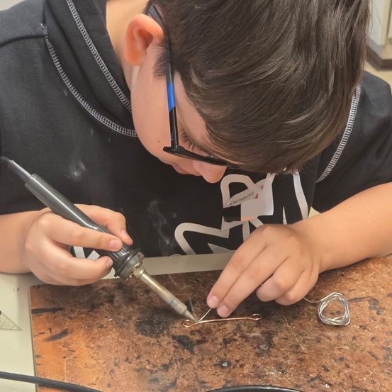 Ein Junge arbeitet mit einem Lötkolben an einem Metallprojekt auf einem Holztisch.