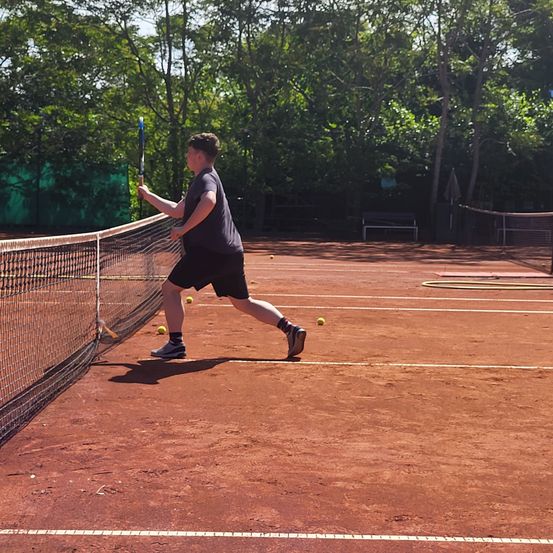 Ein Mann trainiert Tennis auf einem roten Sandplatz, hält einen Schläger und bereitet sich darauf vor, einen Ball in der Nähe des Netzes zu schlagen.