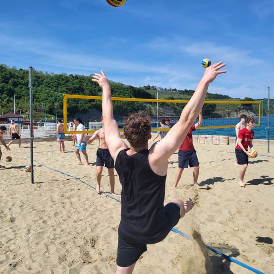An einem Strand spielt eine Gruppe von Menschen Beachvolleyball. Ein Spieler springt in die Luft, um den Ball über das Netz zu schlagen.