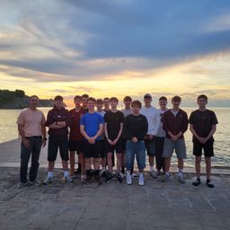 Eine Gruppe junger Männer posiert für ein Foto auf einem Pier, mit Meer und Sonnenuntergang im Hintergrund.