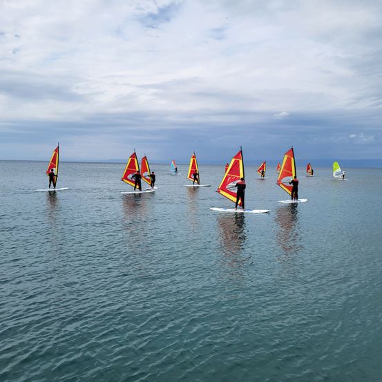 Eine Gruppe von Menschen windsurft auf dem ruhigen Meer, jeder mit einer rot-gelben Segel, unter einem bewölkten Himmel.