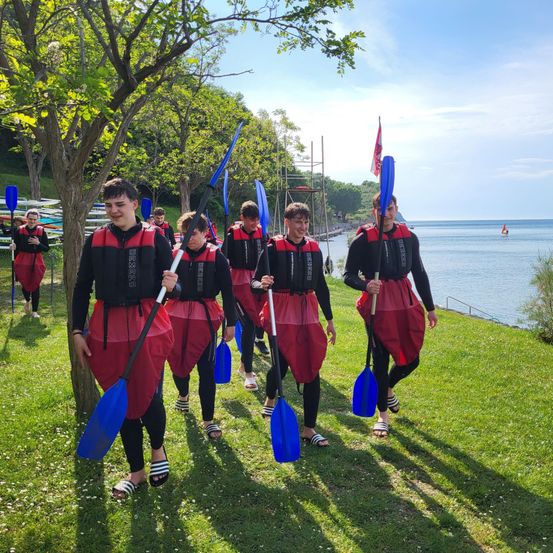 Eine Gruppe junger Leute in roten und schwarzen Schwimmwesten trägt Paddel auf einer Rasenfläche in der Nähe des Wassers.