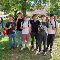 Eine Gruppe junger Männer steht in einem Park, alle halten Snacks in den Händen. Einer trägt eine Fox-Kappe, und ein anderer trägt eine Sonnenbrille. Sie stehen zwischen Bäumen und einem Gebäude im Hintergrund.