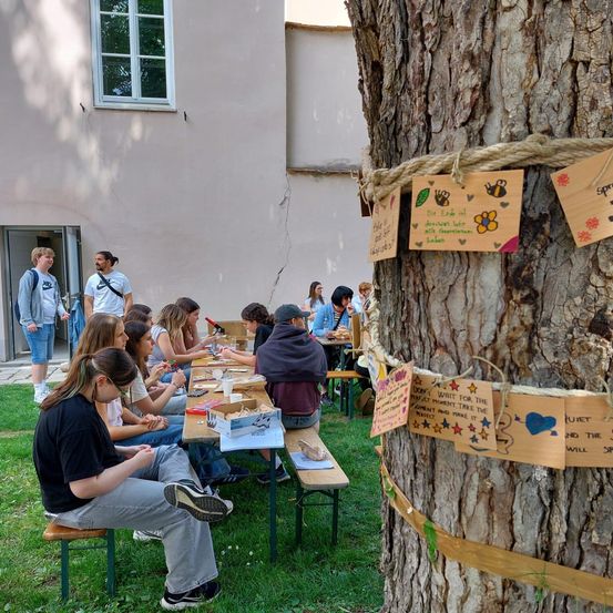Eine Gruppe von Menschen sitzt um Tische in einem Park, einige stehen in der Nähe eines Baumes mit befestigten Notizen. Ein Gebäude mit einem Fenster ist im Hintergrund sichtbar.