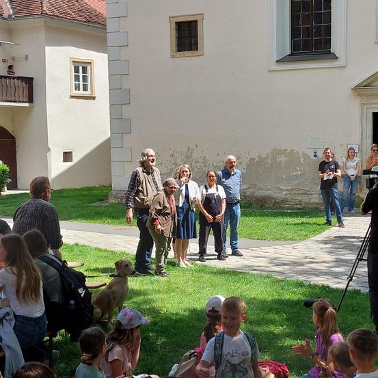 Eine Gruppe von Menschen, darunter Erwachsene und Kinder, versammelt sich auf einem Innenhof. Einige stehen, andere sitzen auf dem Rasen. Ein erwachsener Mann hält einen Hund. Hinter ihnen sind Gebäude mit Fenstern und einem Balkon zu sehen.