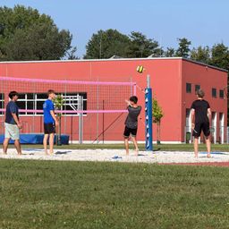 Vier Männer spielen Volleyball auf einem Sandplatz vor einem roten Gebäude. Ein Spieler bereitet sich darauf vor, den Ball zu schlagen.