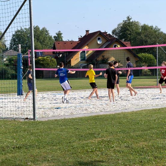 Eine Gruppe von Menschen spielt Beachvolleyball in einer Freiluftumgebung mit einem Haus im Hintergrund. Das Netz ist pink, und sie tragen verschiedenfarbige Trikots.