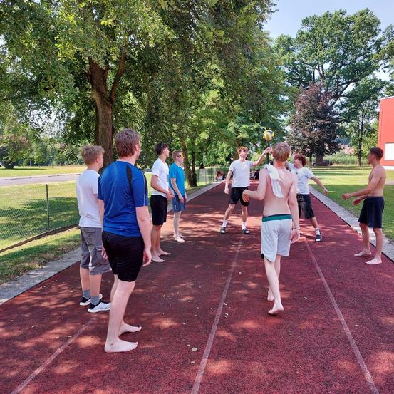 Eine Gruppe junger Männer spielt Volleyball in einem Park auf einer roten Laufbahn mit Bäumen und einem Gebäude im Hintergrund.