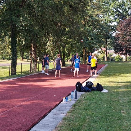 Eine Gruppe von Menschen steht auf einer roten Laufbahn mit Taschen und Flaschen an der Seite. Einige tragen Sportkleidung, und einer ist in der Luft, möglicherweise springend. Bäume und ein Zaun umgeben die Laufbahn.