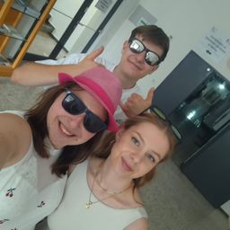 Drei Leute lächeln und posieren für ein Selfie. Ein Mann hinter ihnen gibt einen Daumen hoch. Sie tragen Sonnenbrillen. Ein Glasregal ist hinter dem Mann.