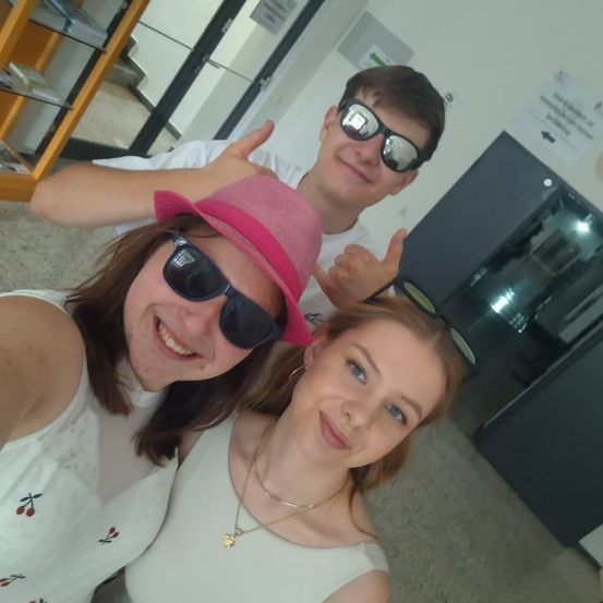 Drei Leute lächeln und posieren für ein Selfie. Ein Mann hinter ihnen gibt einen Daumen hoch. Sie tragen Sonnenbrillen. Ein Glasregal ist hinter dem Mann.