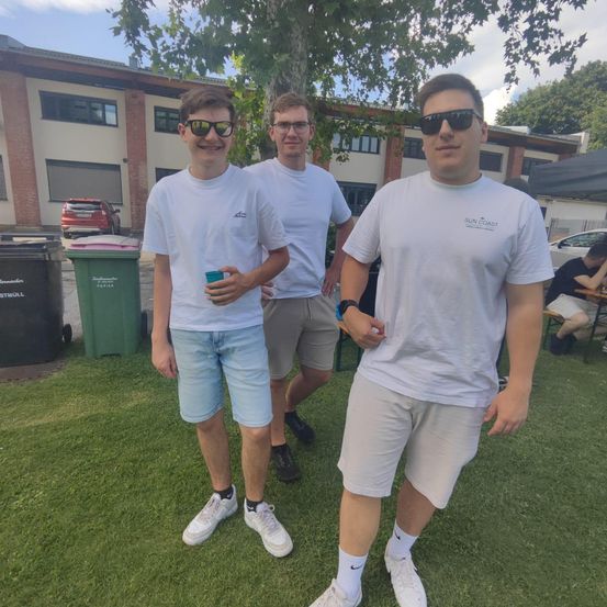 Drei Männer in weißen T-Shirts und Sonnenbrillen stehen auf einem Rasen mit einem Gebäude und Mülleimern im Hintergrund. Einer hält einen grünen Becher.