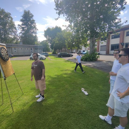Eine Gruppe von Männern spielt Darts auf einem Rasenfeld. Ein Dartboard steht auf einem Staffelei. Ein Mann in Sonnenbrille geht auf dem Bürgersteig.