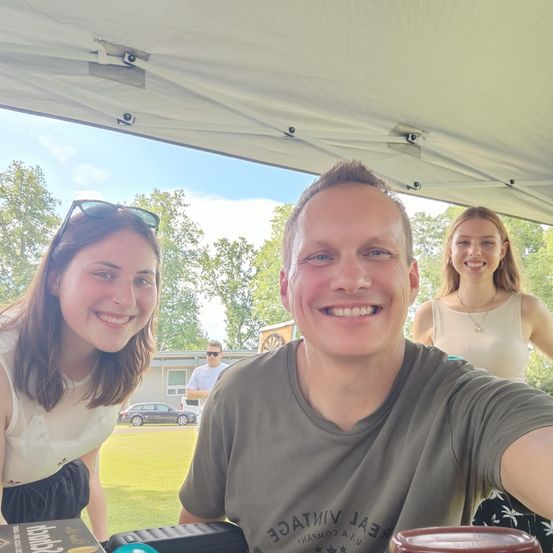 Drei Personen stehen unter einem Zelt und lächeln für ein Selfie. Eine Frau auf der linken Seite trägt eine Sonnenbrille auf dem Kopf, und eine andere Frau steht hinter dem Mann. Ein Auto ist im Hintergrund geparkt.