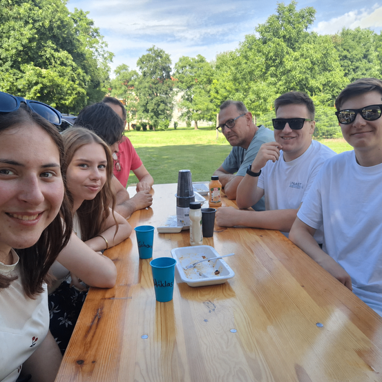 Eine Gruppe von Menschen sitzt an einem Tisch im Park. Einige tragen Sonnenbrillen und alle lächeln. Ein paar Tassen, eine Flasche und ein Tablett mit einem Löffel stehen auf dem Tisch. Bäume und eine Wiese sind im Hintergrund.