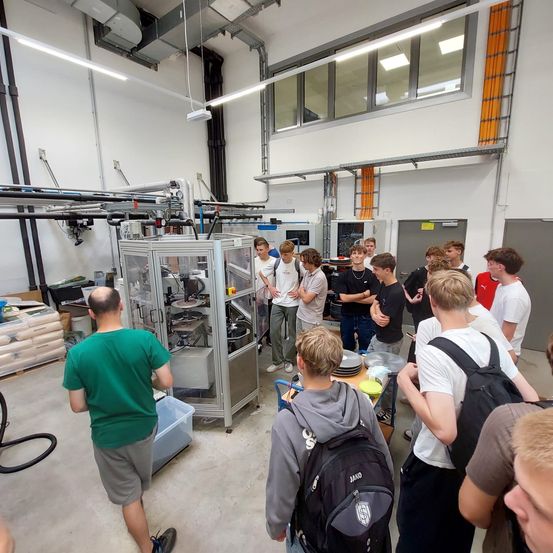 Eine Gruppe von Studenten in einer Werkstatt, die eine große Maschine betrachten, während ein Mann in einem grünen Shirt in der Nähe geht.