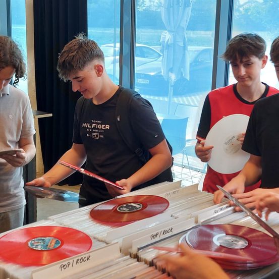 Vier Jungen betrachten Schallplatten. Einer hält eine Platte, während ein anderer eine Schere hält. Sie sind drinnen mit großen Fenstern hinter ihnen.