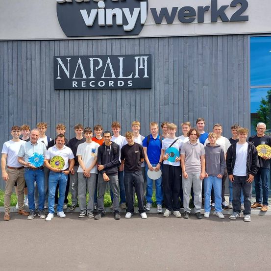 Eine Gruppe von Personen steht vor einem Gebäude mit einem Schild, auf dem 'Vinyl Werk2 Napalm Records' steht. Einige halten kreisförmige Gegenstände und sind lässig gekleidet, einige tragen Hüte.