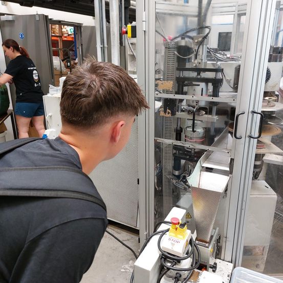 Ein Mann in einer Werkstatt beobachtet eine Maschine, während eine Frau in einem schwarzen Shirt und Shorts in der Nähe arbeitet. Die Maschine hat eine durchsichtige Tür und einen STOP-Knopf.