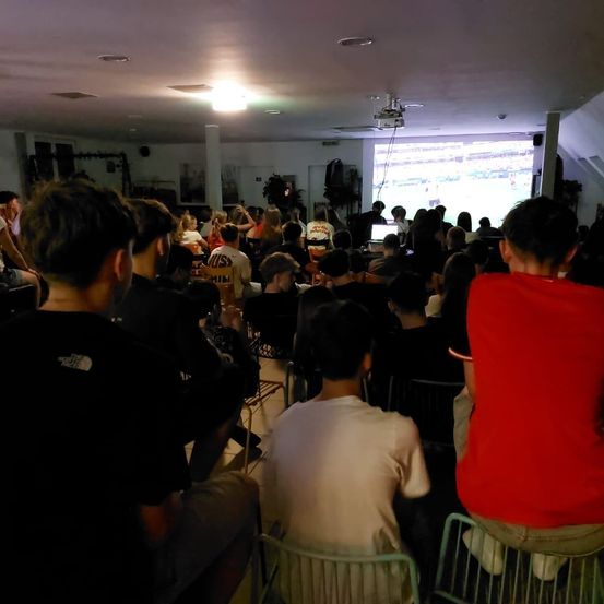 Ein überfüllter Raum mit sitzenden Menschen, die ein Fußballspiel auf einer Projektionsleinwand ansehen. Einige tragen Trikots, andere Freizeitkleidung. Der Raum ist schwach beleuchtet, mit einem Projektor, der von der Decke hängt.