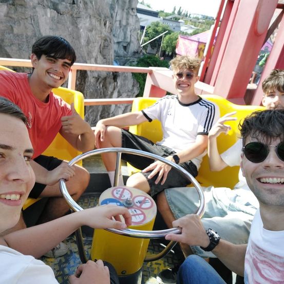 Fünf junge Männer sitzen auf einer Achterbahn in einem Vergnügungspark. Sie lächeln und scheinen für ein Foto zu posieren. Die Achterbahn hat eine gelbe Basis und ein silbernes Lenkrad. Hinter ihnen ist eine Felswand.